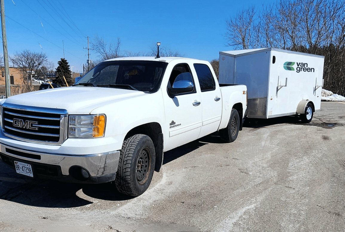 A white GMC pickup truck, marked with the "Lawn Care Hamilton" logo, is towing a white trailer parked on an asphalt surface with patches of snow and bare trees in the background, indicating a